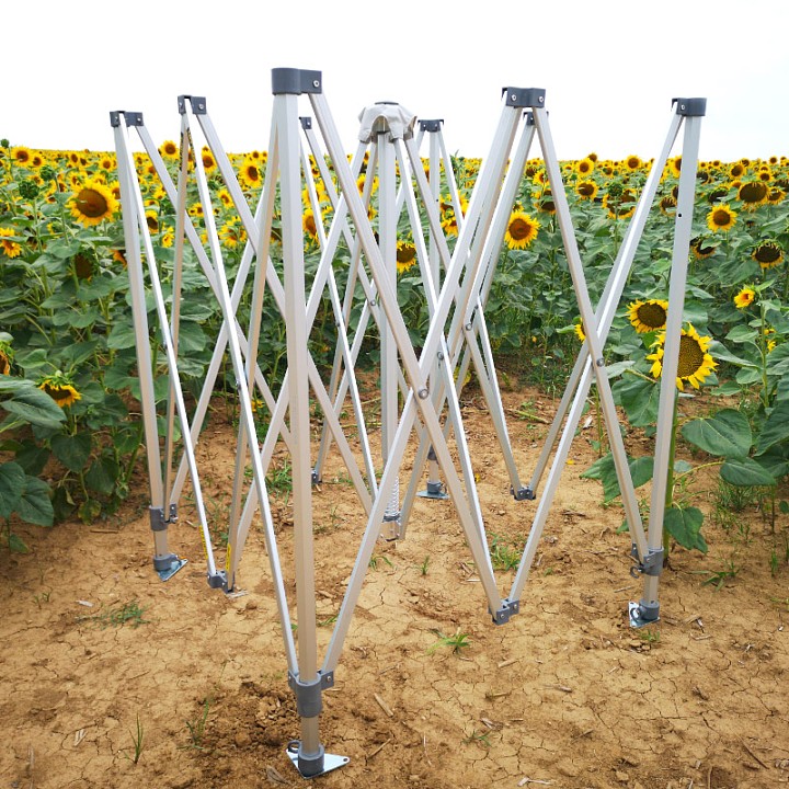 Constructie in een zonnebloemveld: Zilveren schaarframe van een tent uitgeklapt in het veld, dat de stabiliteit van de constructie ook op oneffen, natuurlijke ondergrond laat zien.